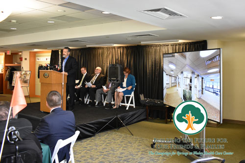a man in a blue suit and shirt presenting to an audience that is out of view. A panel of people sit behind him on a small stage. A screen with a virtual tour of Wesley Health Care Center is in the foreground.