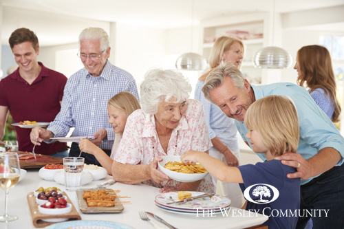 A multi-generational family enjoying dinnertime fun at a kitchen counter. The eldest woman is offering a male child some snacks.
