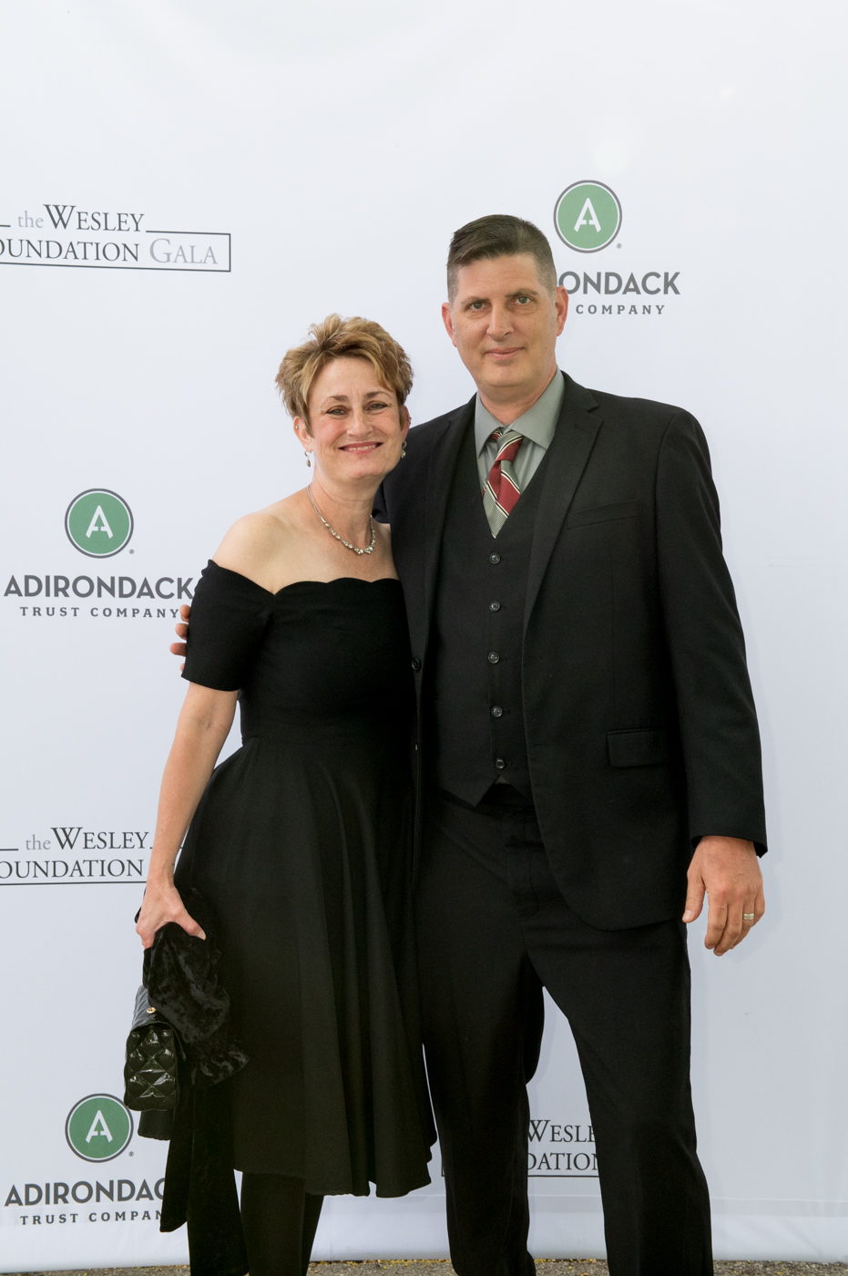 A couple posing for a picture, a woman on the left wearing a black dress and a man wearing a black suit on the right.