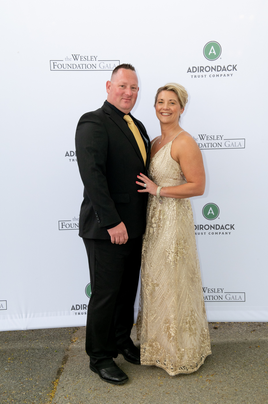 A couple posing for a picture, a man on the left wearing a black suit and a woman on the right wearing a golden dress