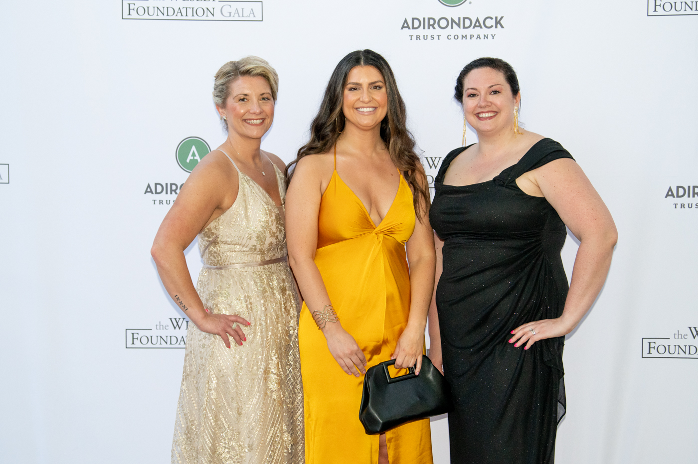 A set of three women posing, from left to right wearing a gold dress, a gold dress and a black dress
